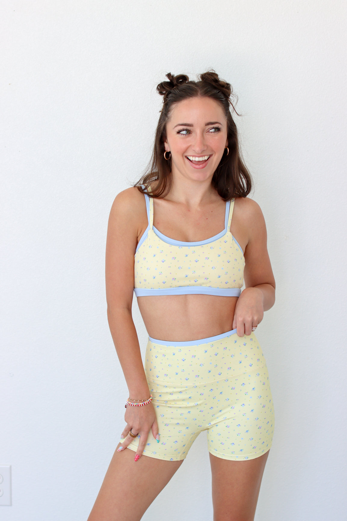 A woman modeling a light yellow two-piece workout set with a small blue floral pattern and light blue trim, consisting of a crop top and matching shorts, against a plain white wall.