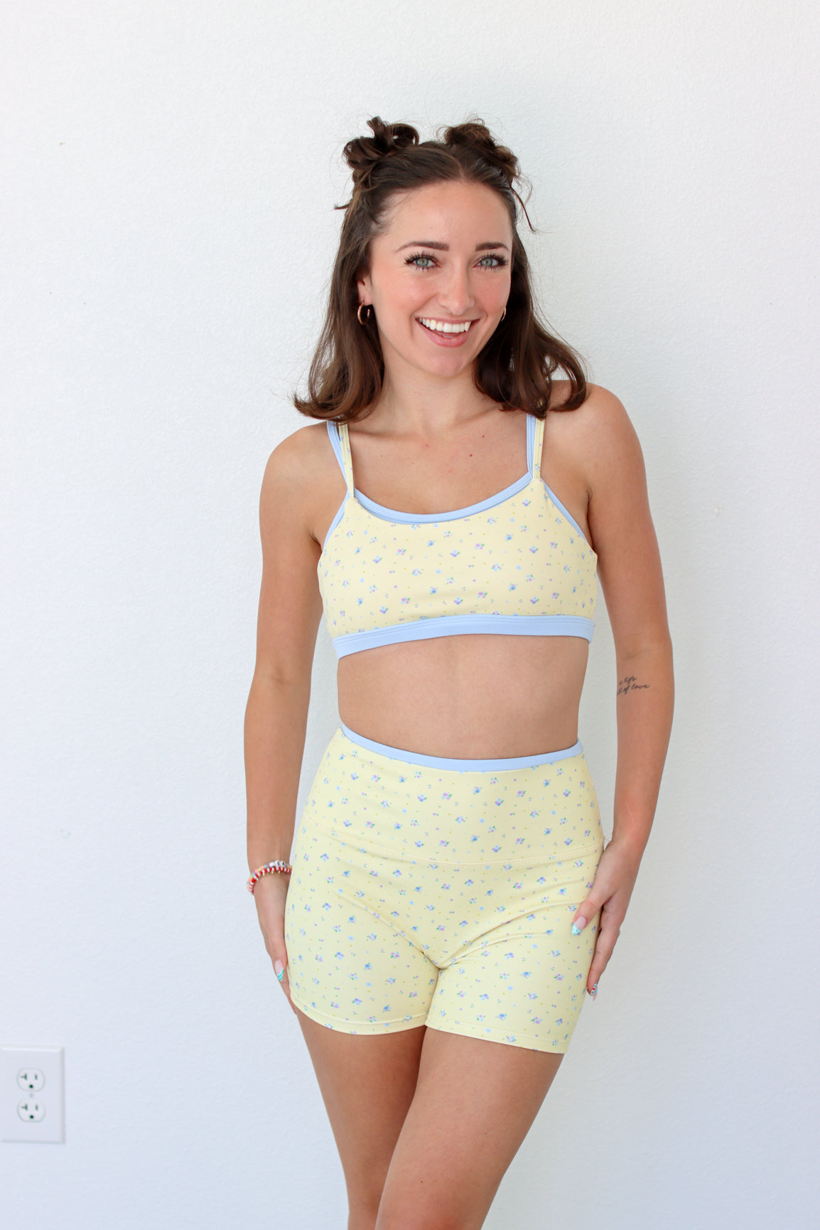 A woman modeling a light yellow two-piece workout set with a small blue floral pattern and light blue trim, consisting of a crop top and matching shorts, against a plain white wall.