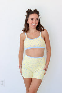 A woman modeling a light yellow two-piece workout set with a small blue floral pattern and light blue trim, consisting of a crop top and matching shorts, against a plain white wall.