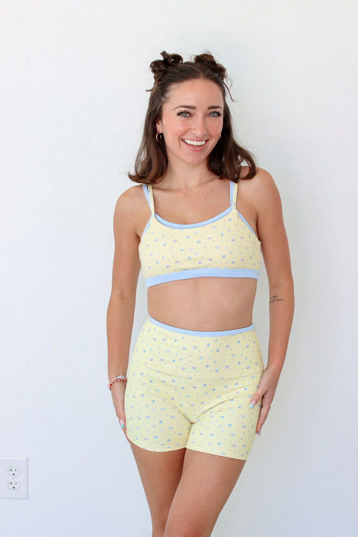 A woman modeling a light yellow two-piece workout set with a small blue floral pattern and light blue trim, consisting of a crop top and matching shorts, against a plain white wall.