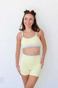 A woman modeling a light yellow two-piece workout set with a small blue floral pattern and light blue trim, consisting of a crop top and matching shorts, against a plain white wall.