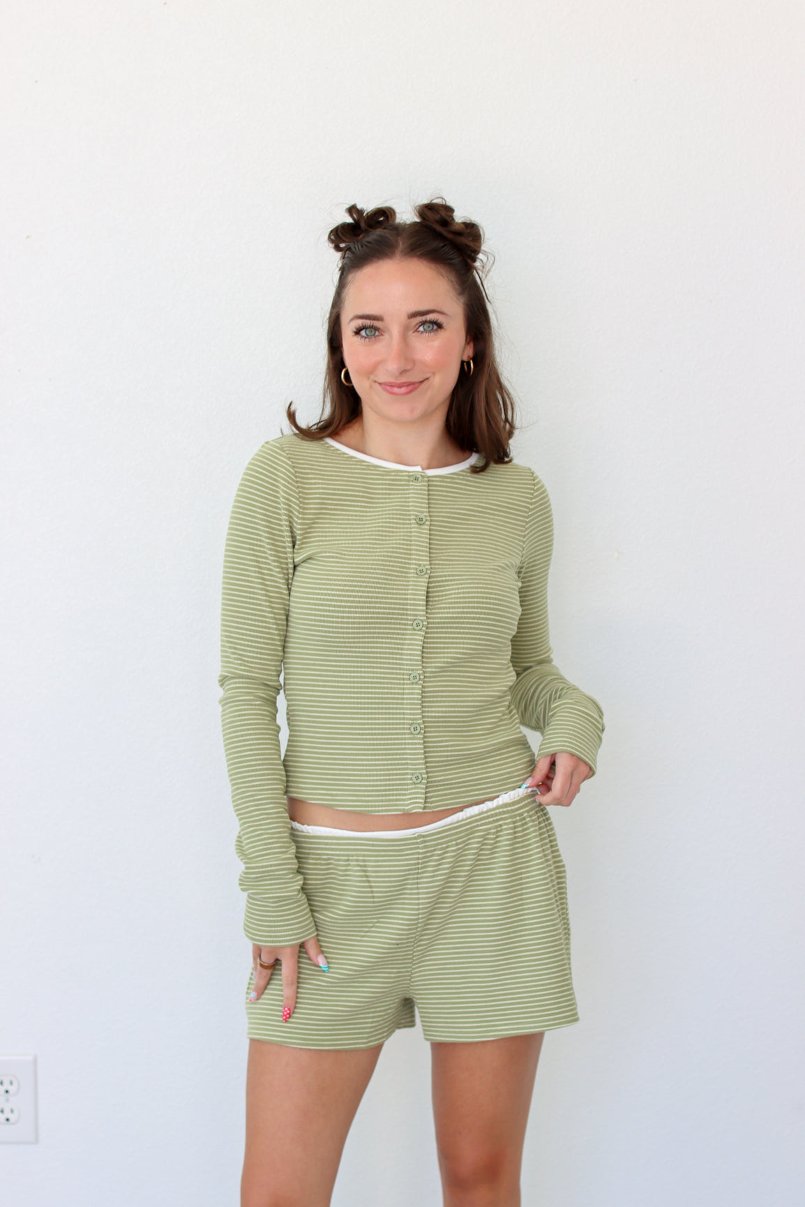 A woman modeling a matching two-piece loungewear set consisting of a long-sleeve button-front top and shorts in a light sage green and white stripe pattern, set against a plain white wall.