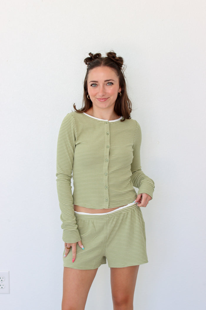 A woman modeling a matching two-piece loungewear set consisting of a long-sleeve button-front top and shorts in a light sage green and white stripe pattern, set against a plain white wall.