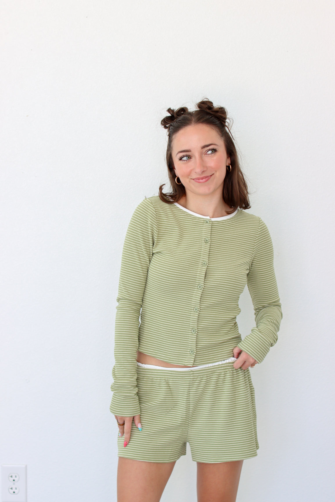 A woman modeling a matching two-piece loungewear set consisting of a long-sleeve button-front top and shorts in a light sage green and white stripe pattern, set against a plain white wall.
