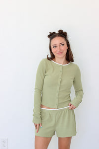 A woman modeling a matching two-piece loungewear set consisting of a long-sleeve button-front top and shorts in a light sage green and white stripe pattern, set against a plain white wall.