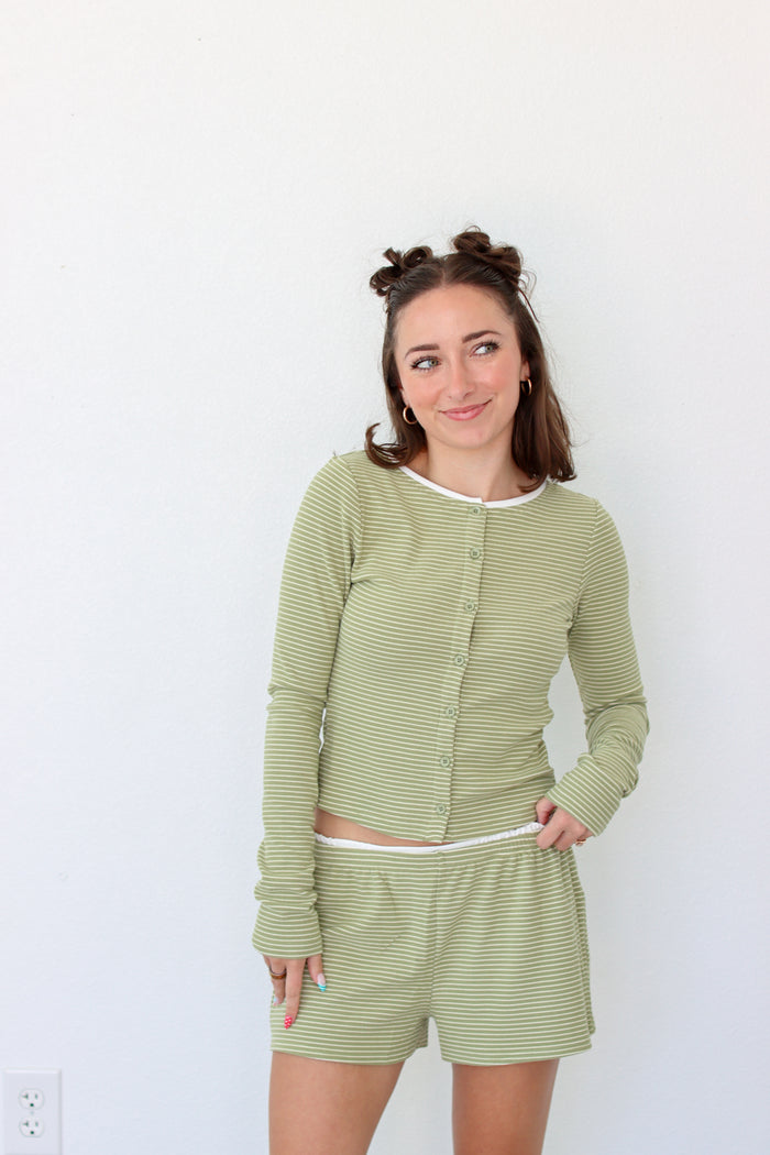 A woman modeling a matching two-piece loungewear set consisting of a long-sleeve button-front top and shorts in a light sage green and white stripe pattern, set against a plain white wall.