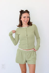 A woman modeling a matching two-piece loungewear set consisting of a long-sleeve button-front top and shorts in a light sage green and white stripe pattern, set against a plain white wall.
