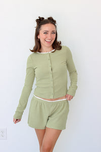 A woman modeling a matching two-piece loungewear set consisting of a long-sleeve button-front top and shorts in a light sage green and white stripe pattern, set against a plain white wall.