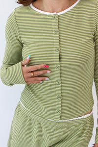 A close-up of a person wearing a light sage green and white striped long-sleeve button-front top and matching bottoms, with the hand resting on the chest.