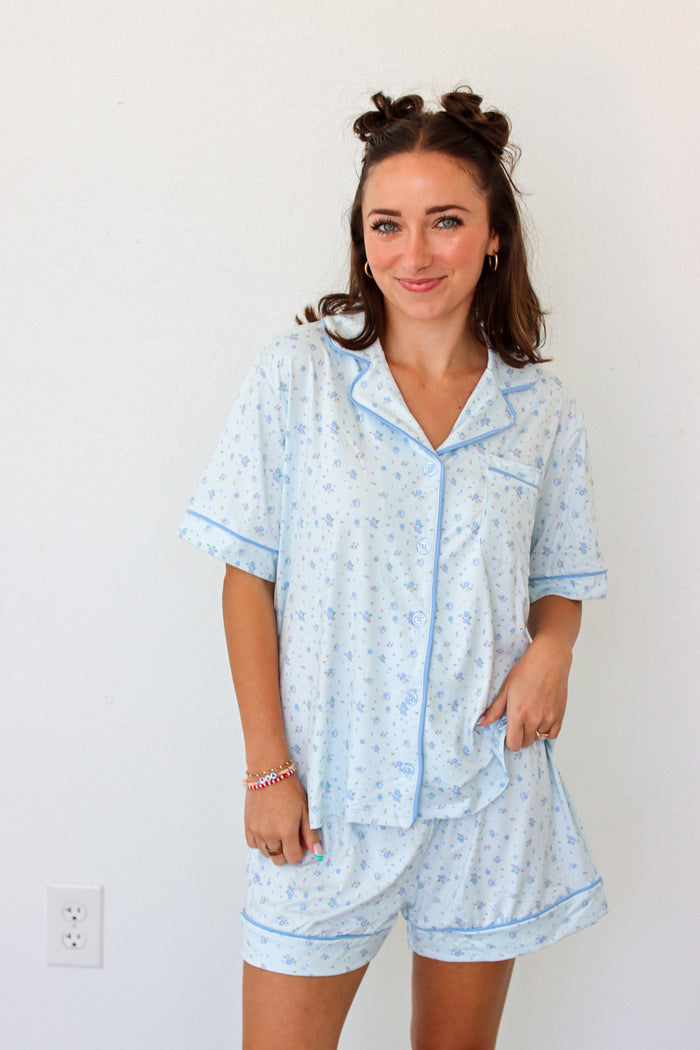 A woman wearing a light blue and white patterned pajama set with matching shorts and a short-sleeve button-front top, standing against a plain white wall.
