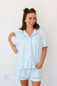 A woman wearing a light blue and white patterned pajama set with matching shorts and a short-sleeve button-front top, standing against a plain white wall.