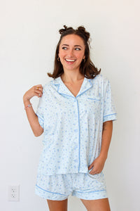 A woman wearing a light blue and white patterned pajama set with matching shorts and a short-sleeve button-front top, standing against a plain white wall.