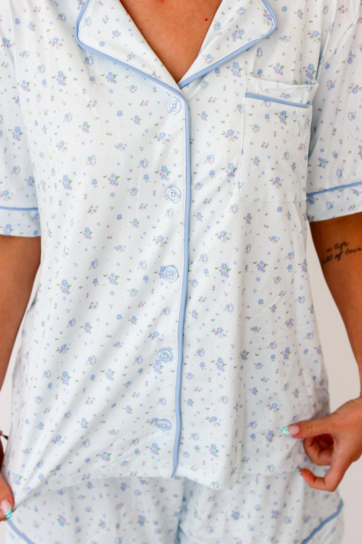 A close-up of a person wearing a short-sleeve pajama top with a light blue base, light blue floral print, and light blue piping trim. The pajama top has a button-down front and a chest pocket.