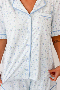 A close-up of a person wearing a short-sleeve pajama top with a light blue base, light blue floral print, and light blue piping trim. The pajama top has a button-down front and a chest pocket.