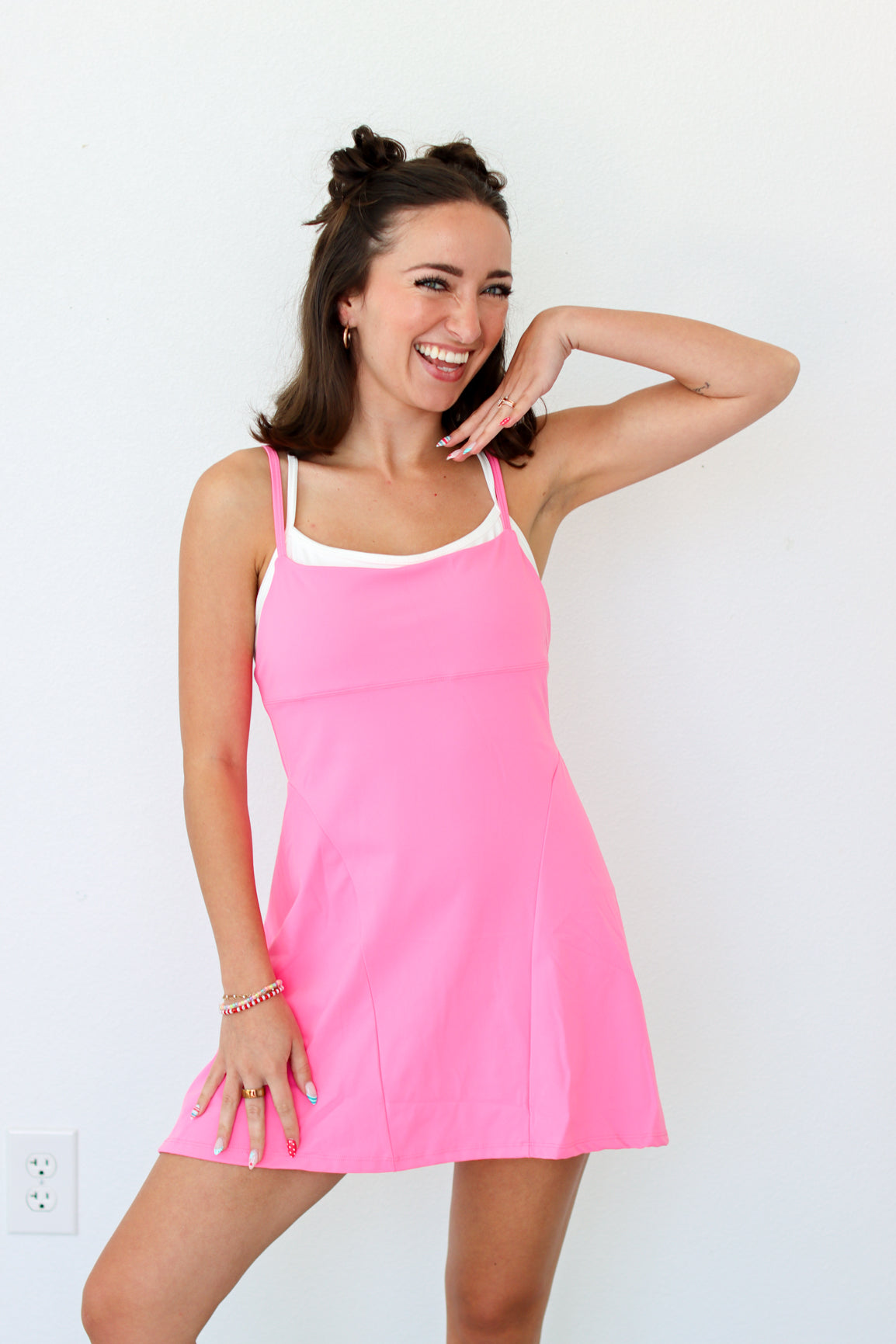 A woman modeling a bright pink sleeveless mini dress with thin straps and a white inner neckline, standing against a plain white wall.