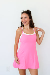 A woman modeling a bright pink sleeveless mini dress with thin straps and a white inner neckline, standing against a plain white wall.