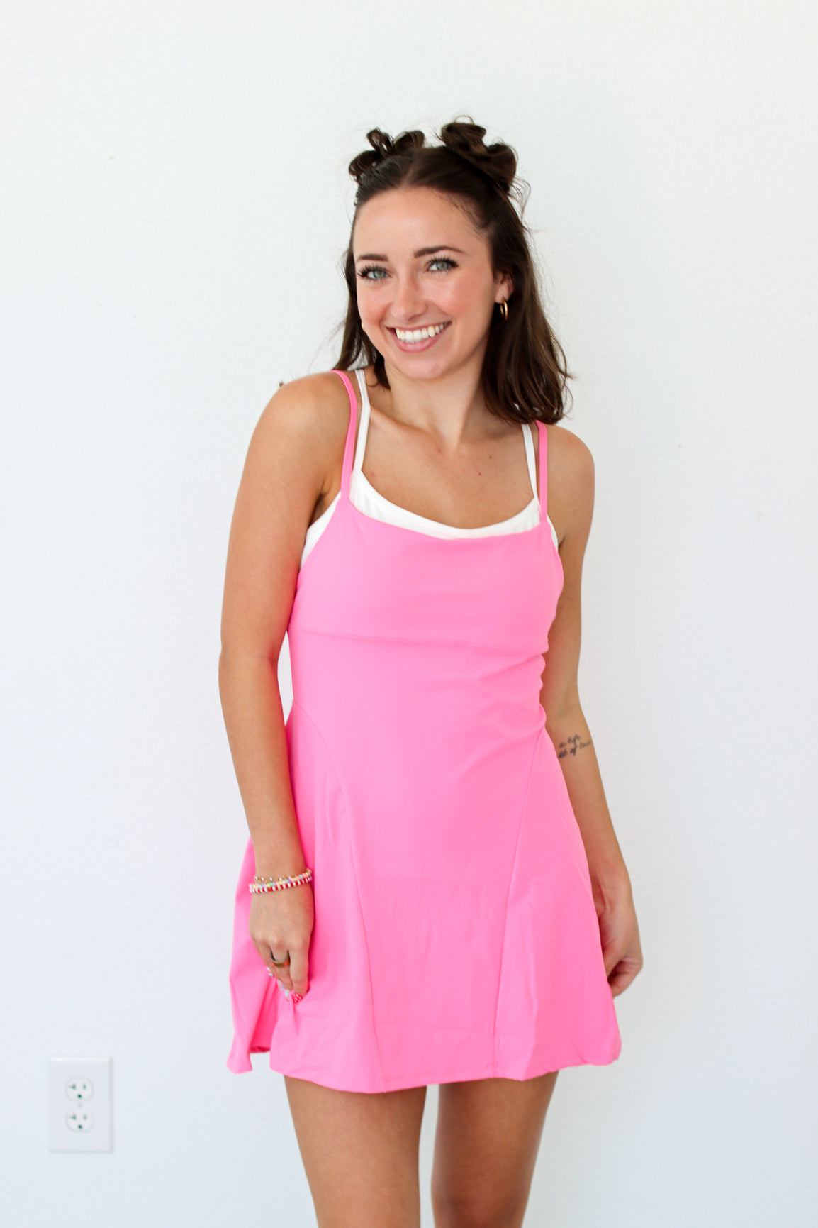 A woman modeling a bright pink sleeveless mini dress with thin straps and a white inner neckline, standing against a plain white wall.