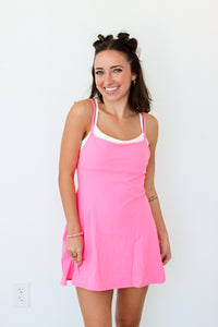 A woman modeling a bright pink sleeveless mini dress with thin straps and a white inner neckline, standing against a plain white wall.