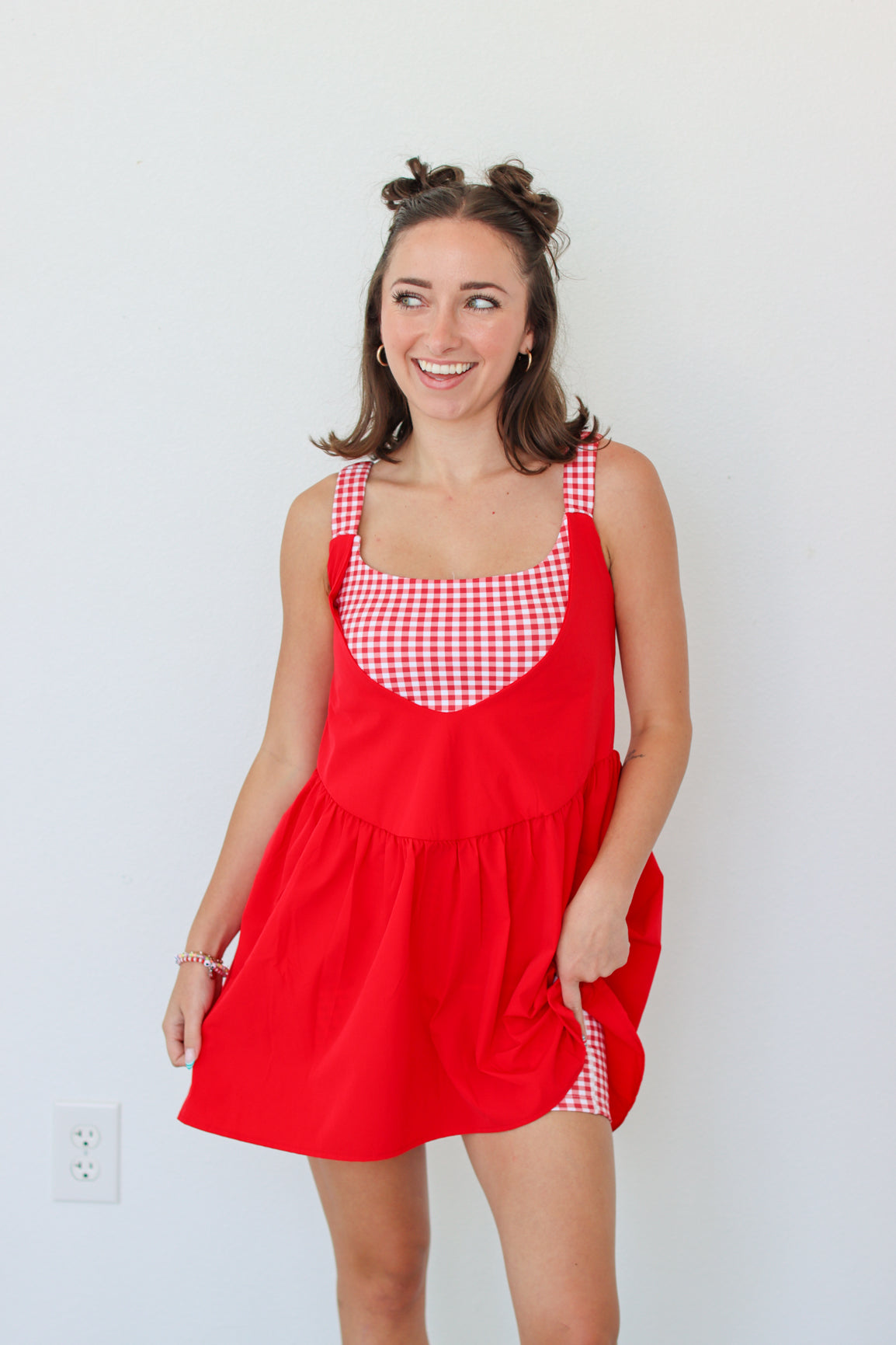 A woman modeling a red pinafore-style dress with red and white gingham trim on the bodice and straps, standing against a plain white wall.