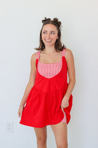 A woman modeling a red pinafore-style dress with red and white gingham trim on the bodice and straps, standing against a plain white wall.