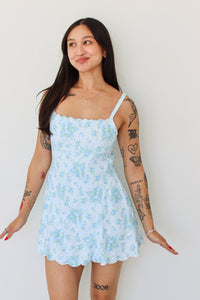 A woman with visible arm tattoos models a light blue and white floral print mini dress with thin straps and a scalloped hem against a plain white wall.