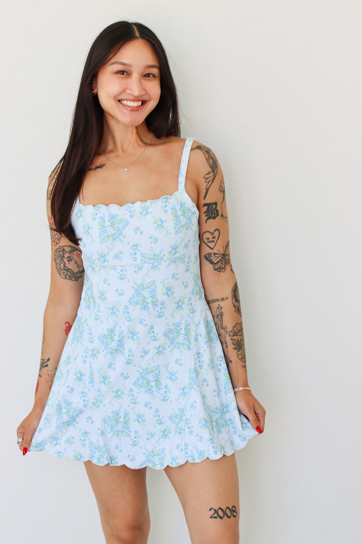 A woman with long dark hair and visible arm tattoos is wearing a light blue and white floral print mini dress with scalloped edges, standing against a plain white wall.