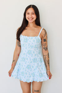 A woman with long dark hair and visible arm tattoos is wearing a light blue and white floral print mini dress with scalloped edges, standing against a plain white wall.