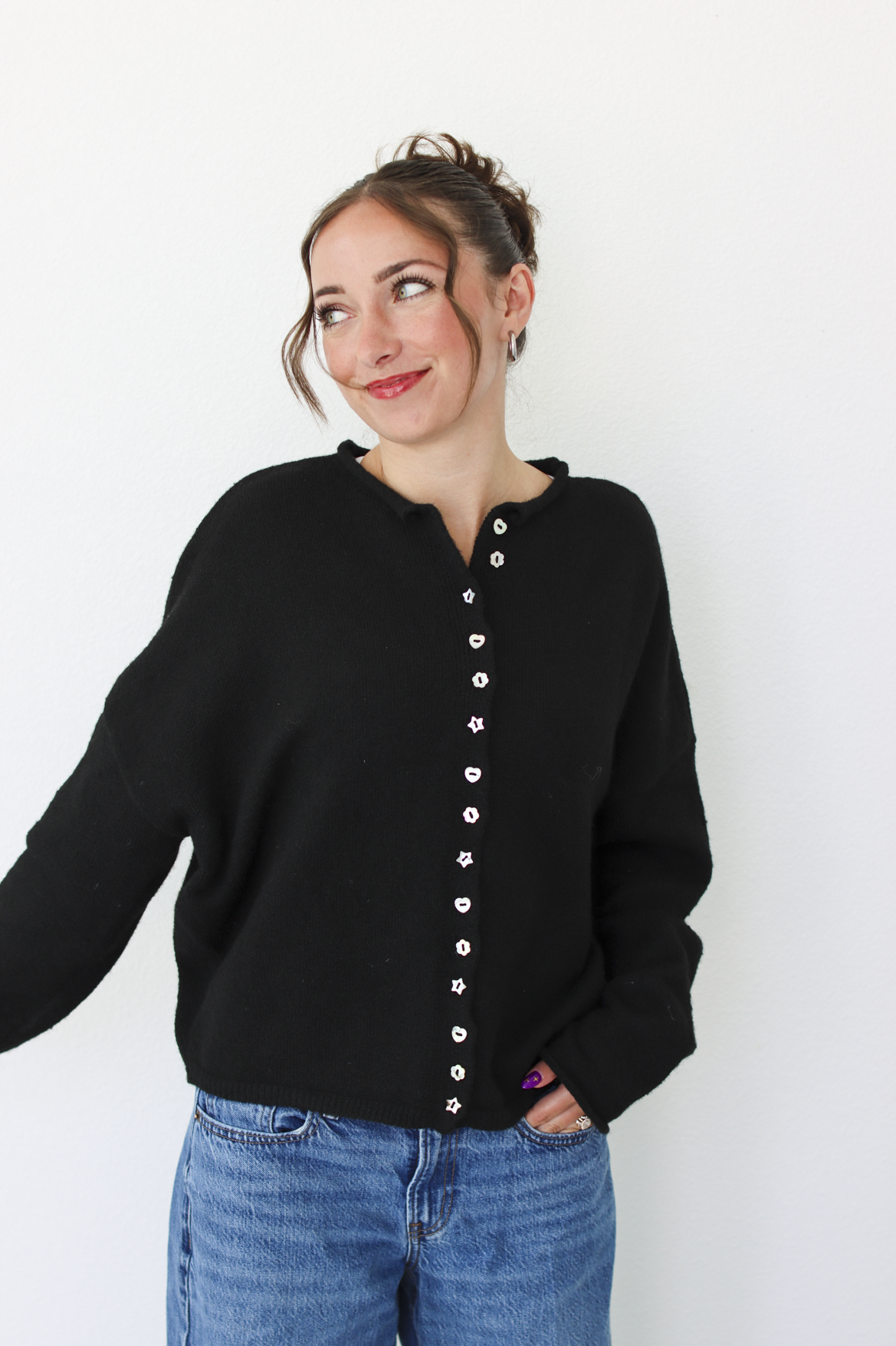 A woman wearing a black knit cardigan sweater with a decorative button-down front and blue jeans, standing against a plain white wall.