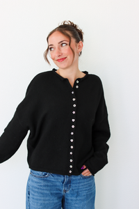 A woman wearing a black knit cardigan sweater with a decorative button-down front and blue jeans, standing against a plain white wall.