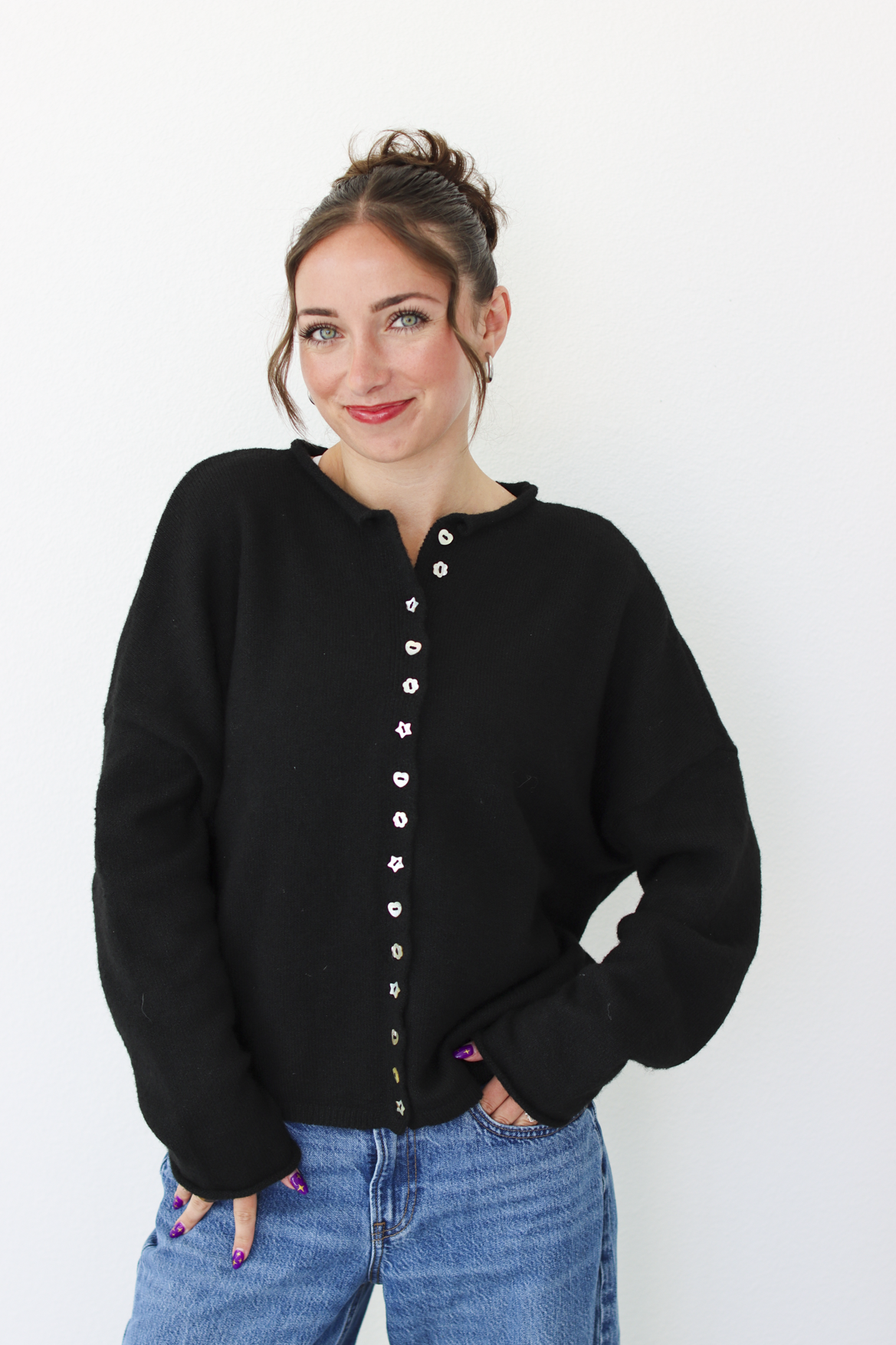 A woman wearing a black knit cardigan sweater with a decorative button-down front and blue jeans, standing against a plain white wall.