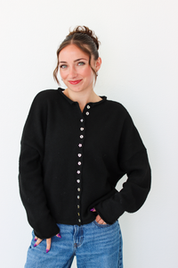 A woman wearing a black knit cardigan sweater with a decorative button-down front and blue jeans, standing against a plain white wall.