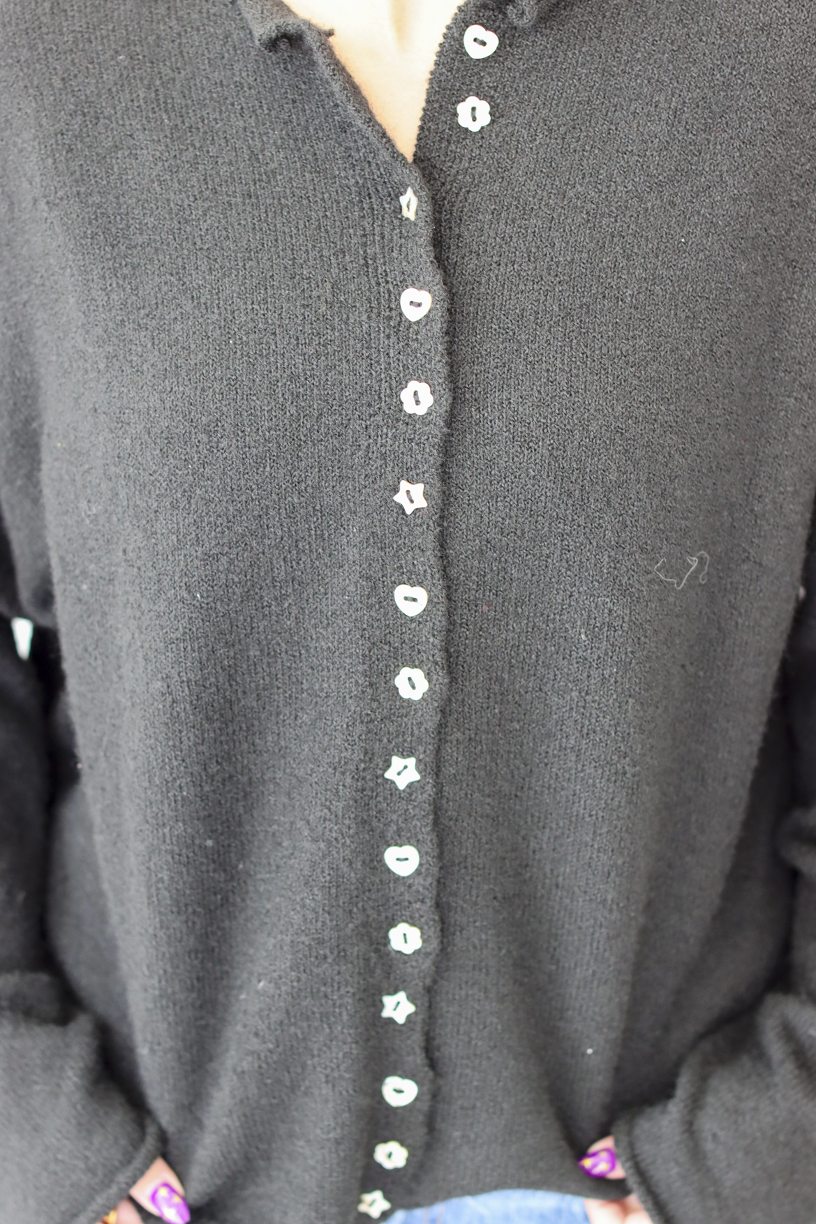 A woman wearing a black knit cardigan sweater with a decorative button-down front and blue jeans, standing against a plain white wall.