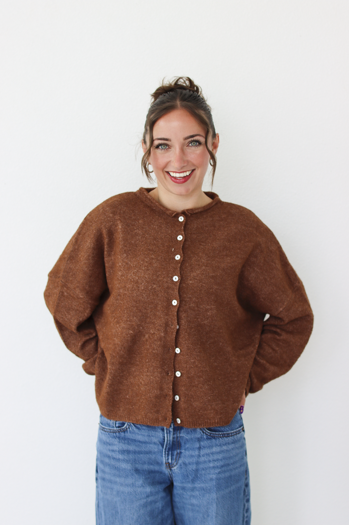 A woman smiling and wearing a brown cardigan with white buttons, paired with blue jeans, standing against a plain white wall.