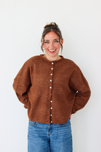 A woman smiling and wearing a brown cardigan with white buttons, paired with blue jeans, standing against a plain white wall.