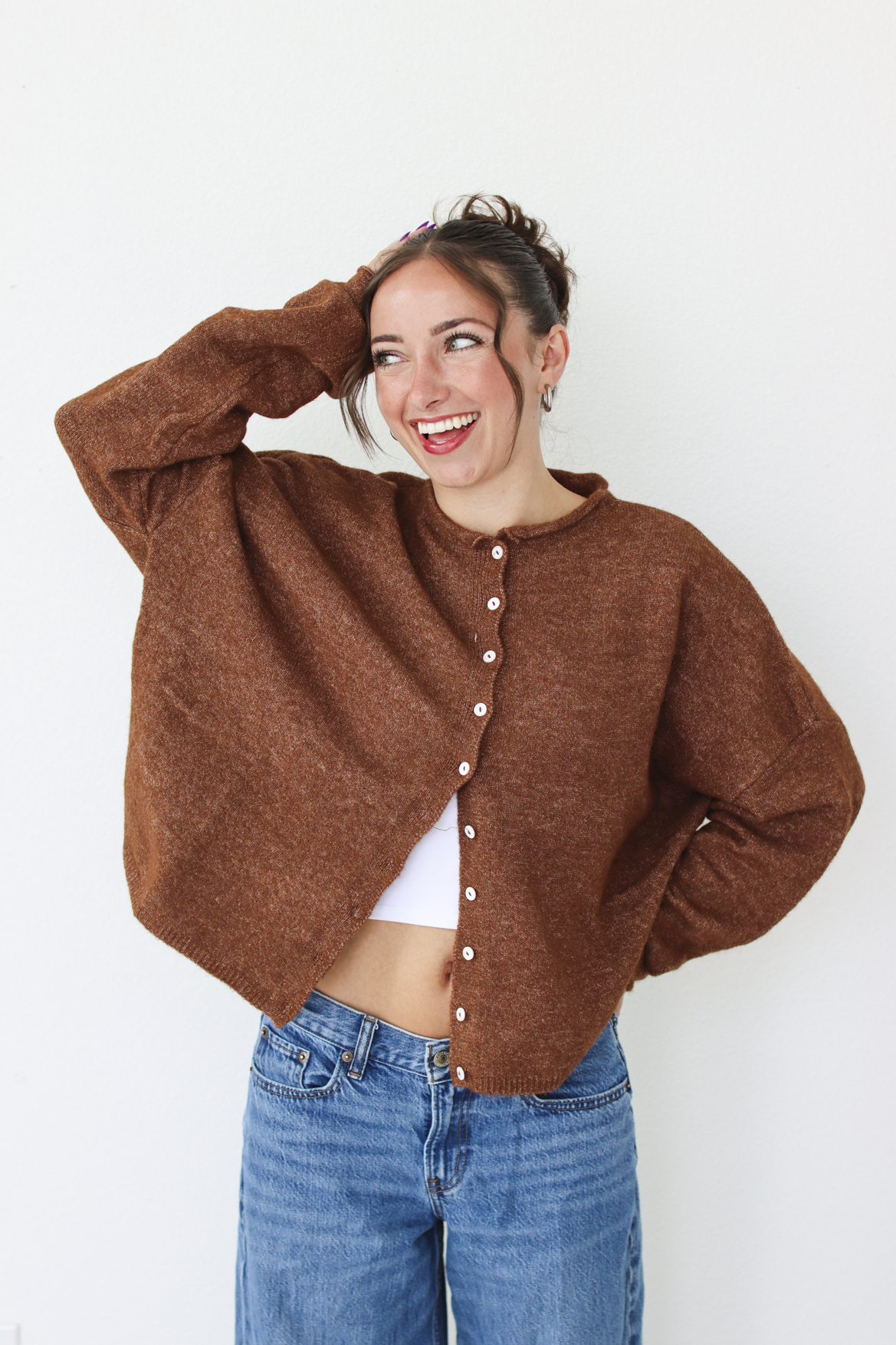 A woman smiling and wearing a brown cardigan with white buttons, paired with blue jeans, standing against a plain white wall.