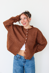 A woman smiling and wearing a brown cardigan with white buttons, paired with blue jeans, standing against a plain white wall.