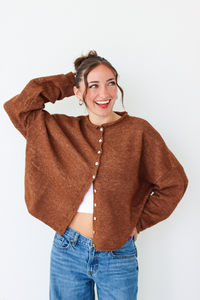 A woman smiling and wearing a brown cardigan with white buttons, paired with blue jeans, standing against a plain white wall.
