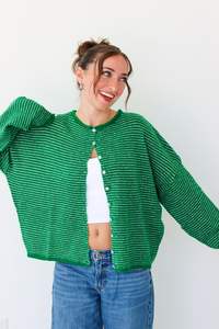 A woman smiling and wearing a bright green striped knit cardigan with a white  top and blue jeans, standing against a plain white wall.