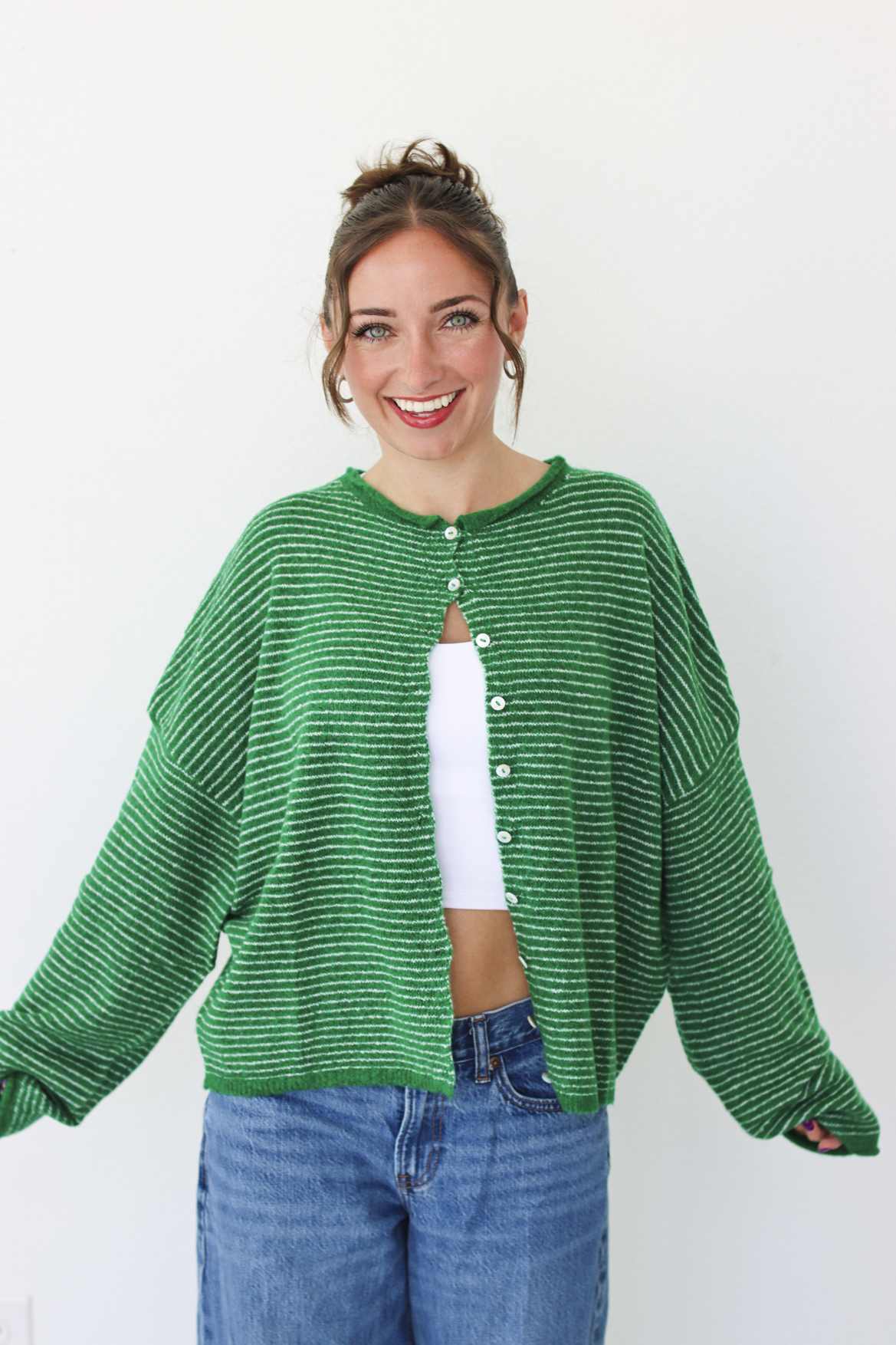 A woman smiling and wearing a bright green striped knit cardigan with a white  top and blue jeans, standing against a plain white wall.