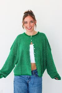 A woman smiling and wearing a bright green striped knit cardigan with a white  top and blue jeans, standing against a plain white wall.