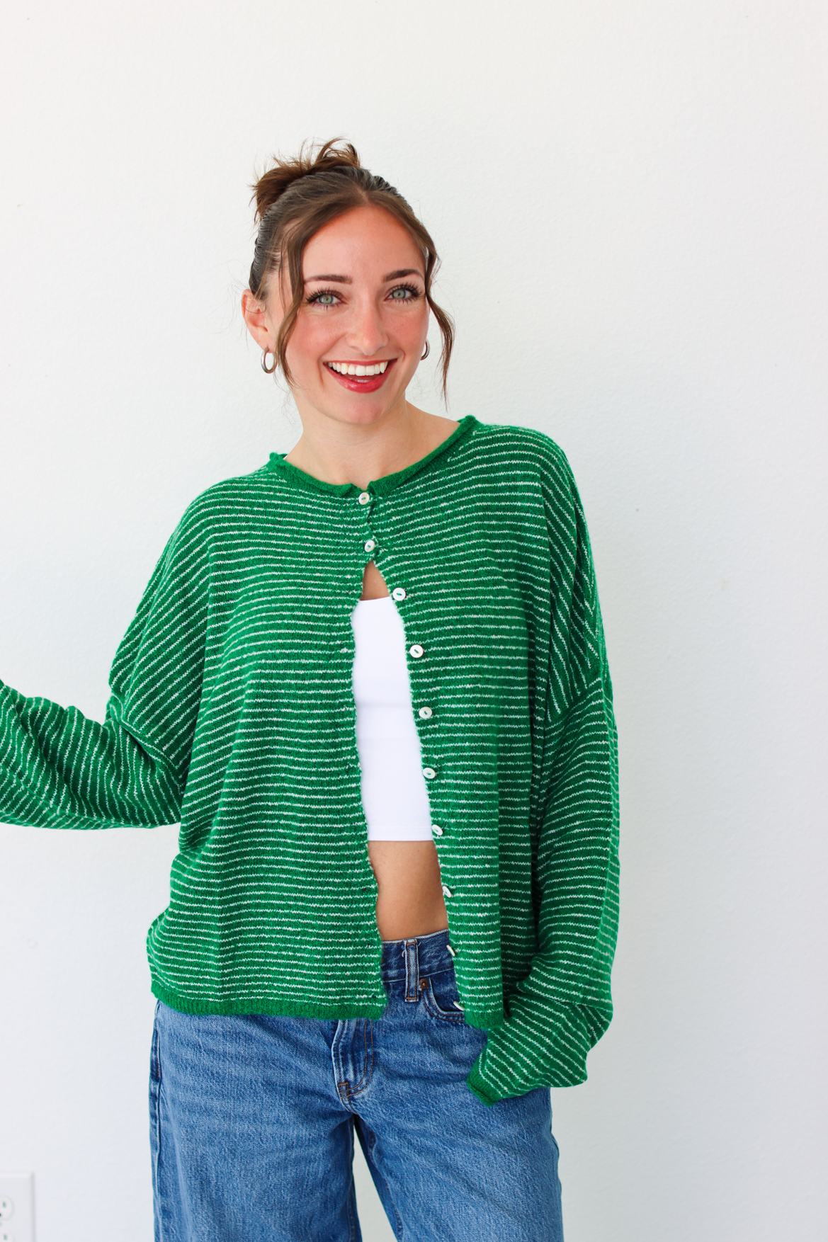 A woman smiling and wearing a bright green striped knit cardigan with a white  top and blue jeans, standing against a plain white wall.