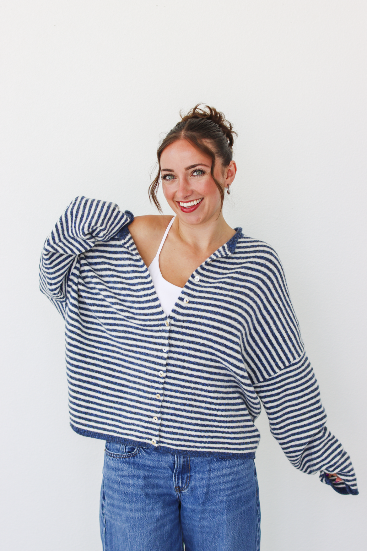 A woman modeling a navy and white striped button-up cardigan over a white tank top and blue jeans, standing against a plain white background.
