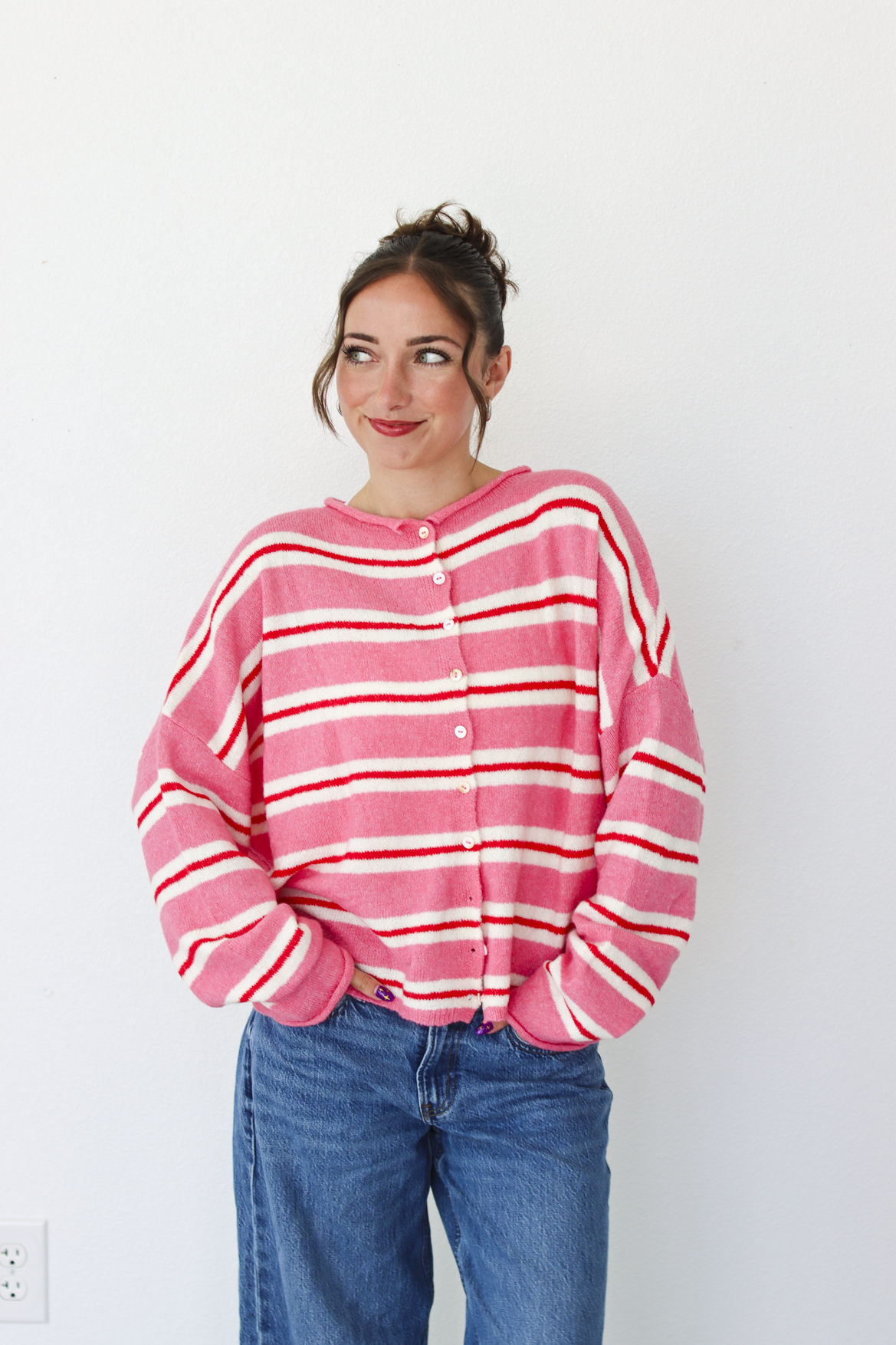 A woman smiling and wearing a pink cardigan with thin red and white horizontal stripes, paired with blue jeans, standing against a plain white wall.