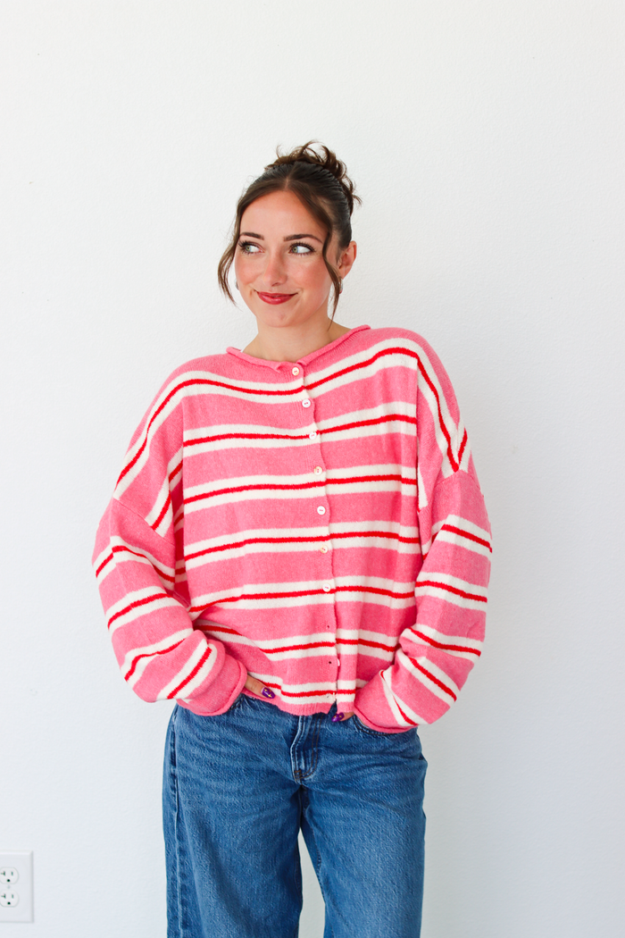 A woman smiling and wearing a pink cardigan with thin red and white horizontal stripes, paired with blue jeans, standing against a plain white wall.