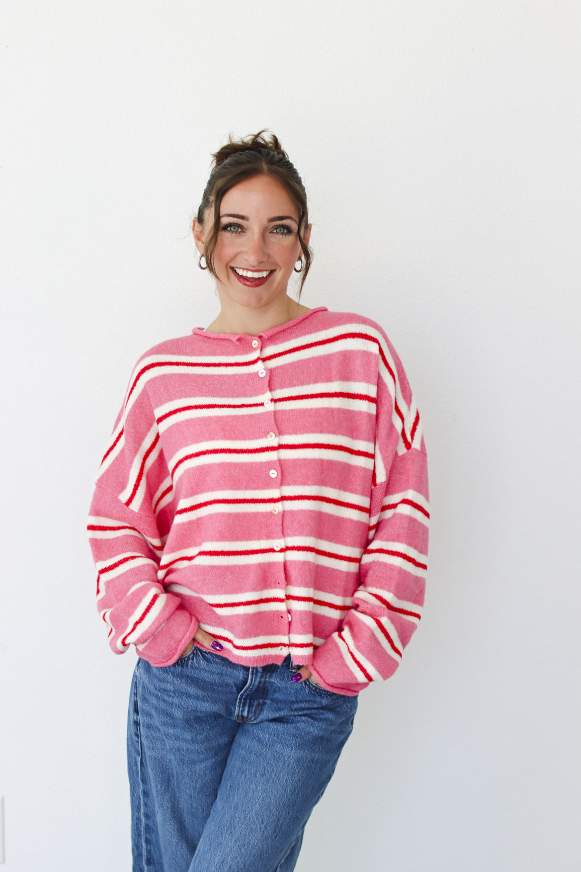 A woman smiling and wearing a pink cardigan with thin red and white horizontal stripes, paired with blue jeans, standing against a plain white wall.