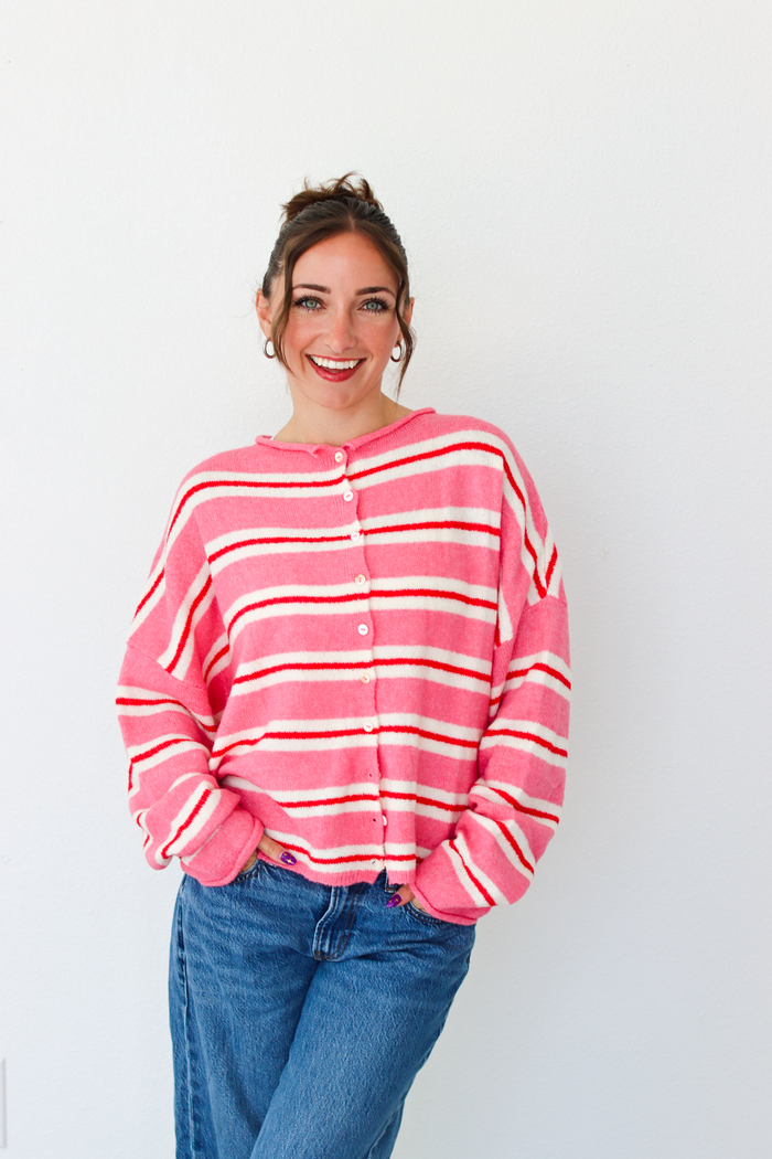 A woman smiling and wearing a pink cardigan with thin red and white horizontal stripes, paired with blue jeans, standing against a plain white wall.