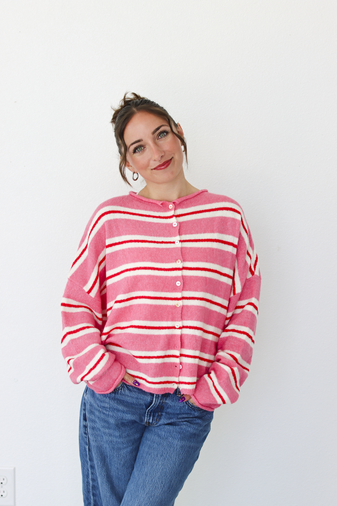 A woman smiling and wearing a pink cardigan with thin red and white horizontal stripes, paired with blue jeans, standing against a plain white wall.
