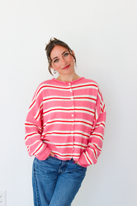 A woman smiling and wearing a pink cardigan with thin red and white horizontal stripes, paired with blue jeans, standing against a plain white wall.
