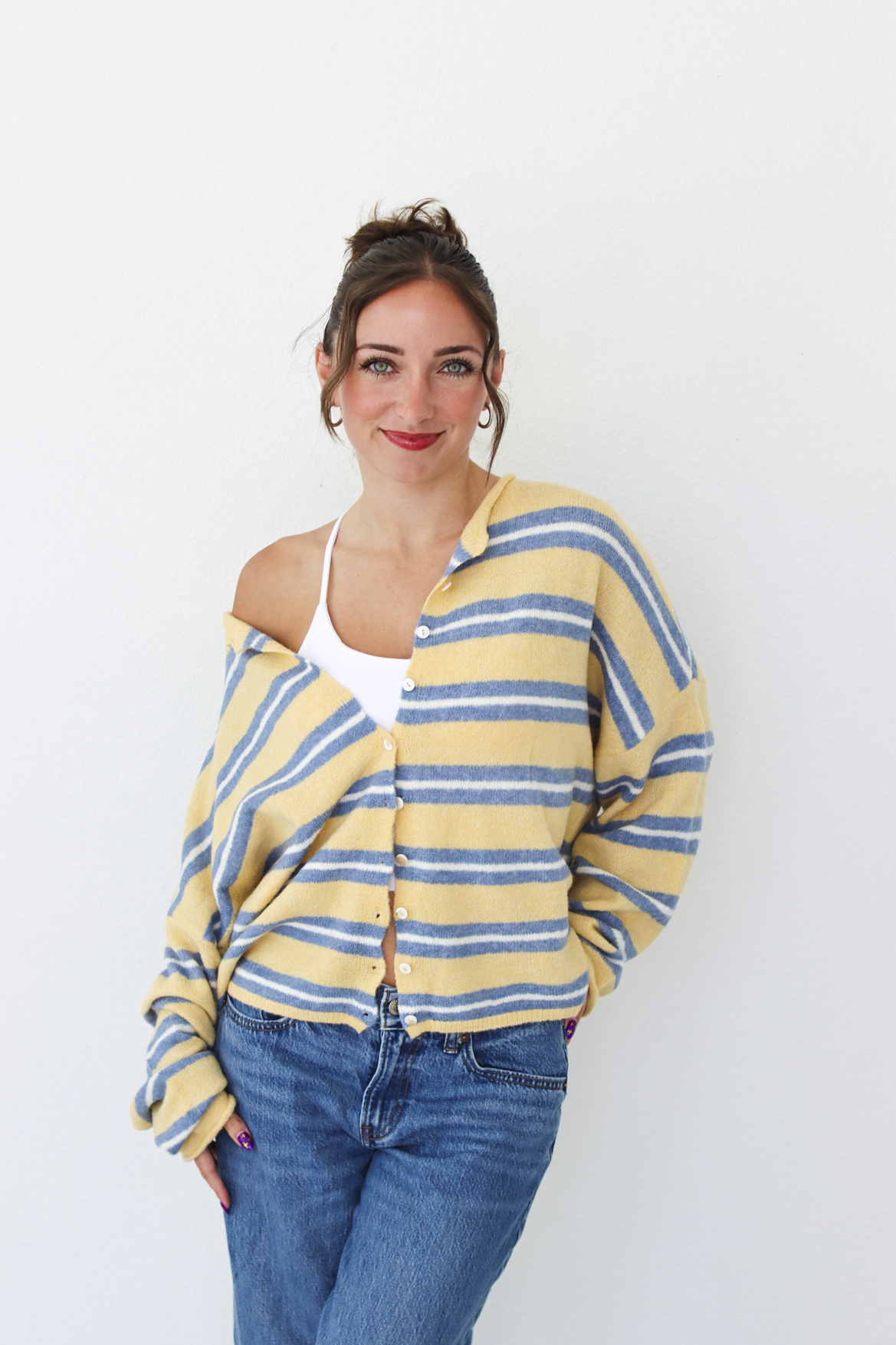 A woman wearing a yellow and blue striped knit cardigan over a white tank top and blue jeans, standing against a plain white wall.