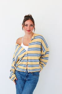 A woman wearing a yellow and blue striped knit cardigan over a white tank top and blue jeans, standing against a plain white wall.
